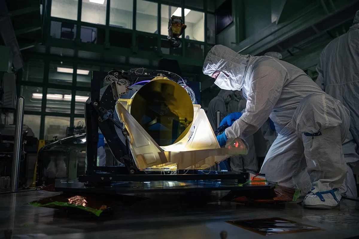 A person in cleanroom clothing works beside the gold-mirrored LISA telescope prototype.