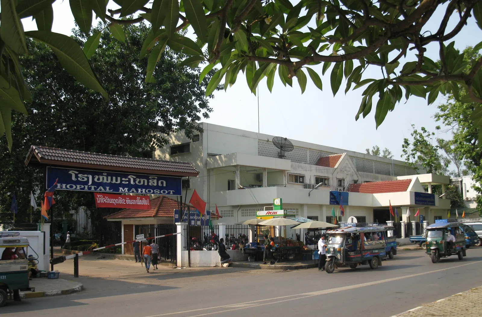 Entrance to Mahosot Hospital in Vientiane, with trees and vehicles on the road