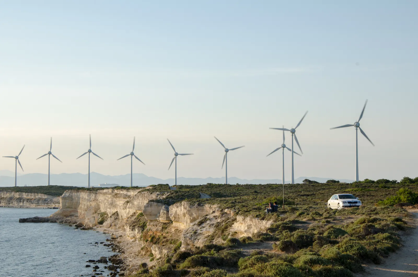 Wind turbines along a rocky coast in Türkiye, with sea and vegetation in the foreground.