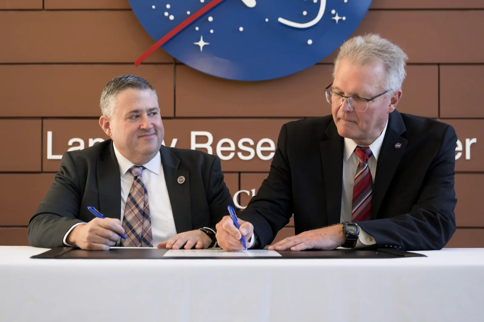 Vince Maiden on the left and Allen Kilgore on the right sign the agreement in front of the NASA Langley emblem.