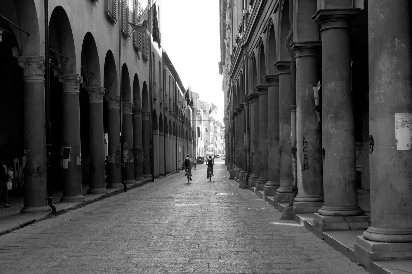 The porticoes of Via Zamboni in Bologna, with bicycles and people along the street, in a black-and-white photograph.