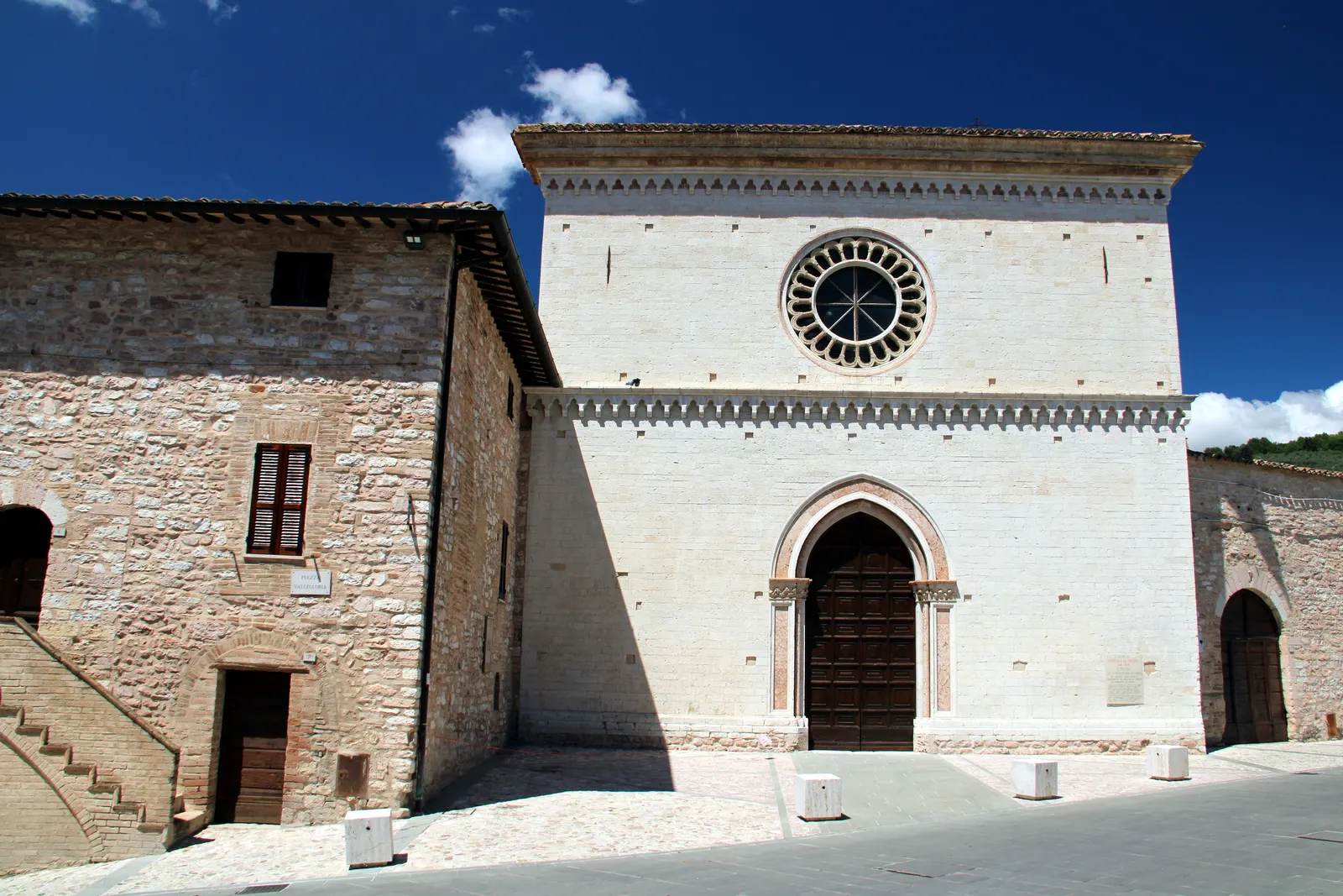 The pale stone facade of Santa Maria di Vallegloria church in Spello, with a rose window and an arched doorway.